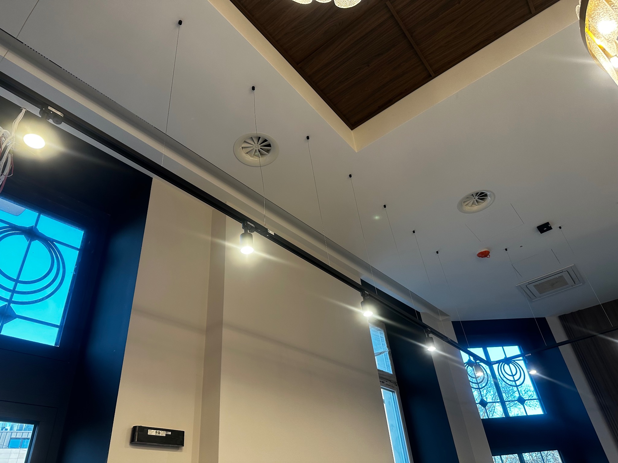 Nowoczesne oświetlenie szynowe z reflektorami punktowymi, widok z dołu na tle okien z dekoracyjnymi szybami i sufitu z elementami drewnianymi.