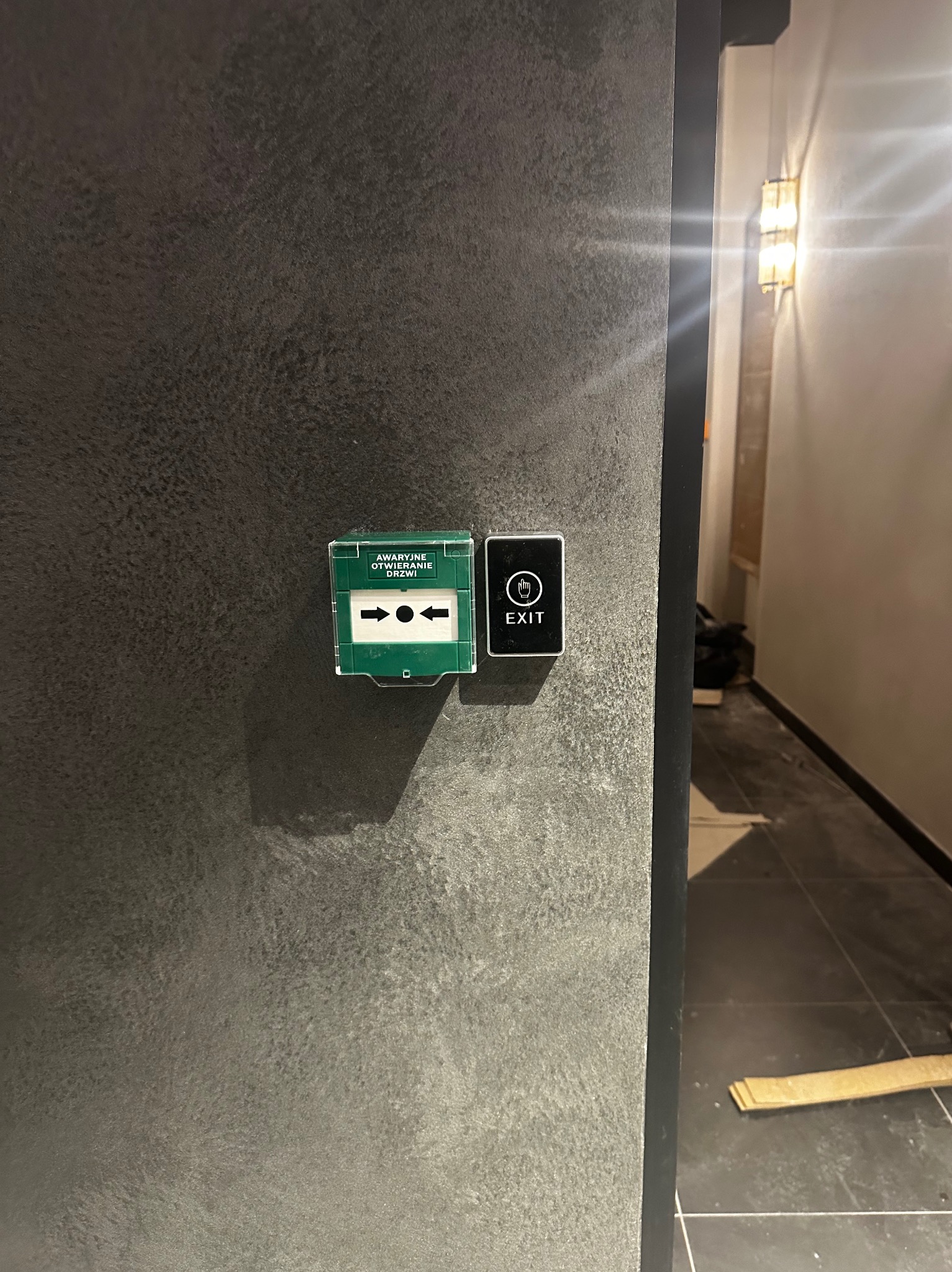 Przycisk awaryjnego otwierania drzwi i panel dotykowy EXIT zamontowane na szarej ścianie w korytarzu z oświetleniem.
