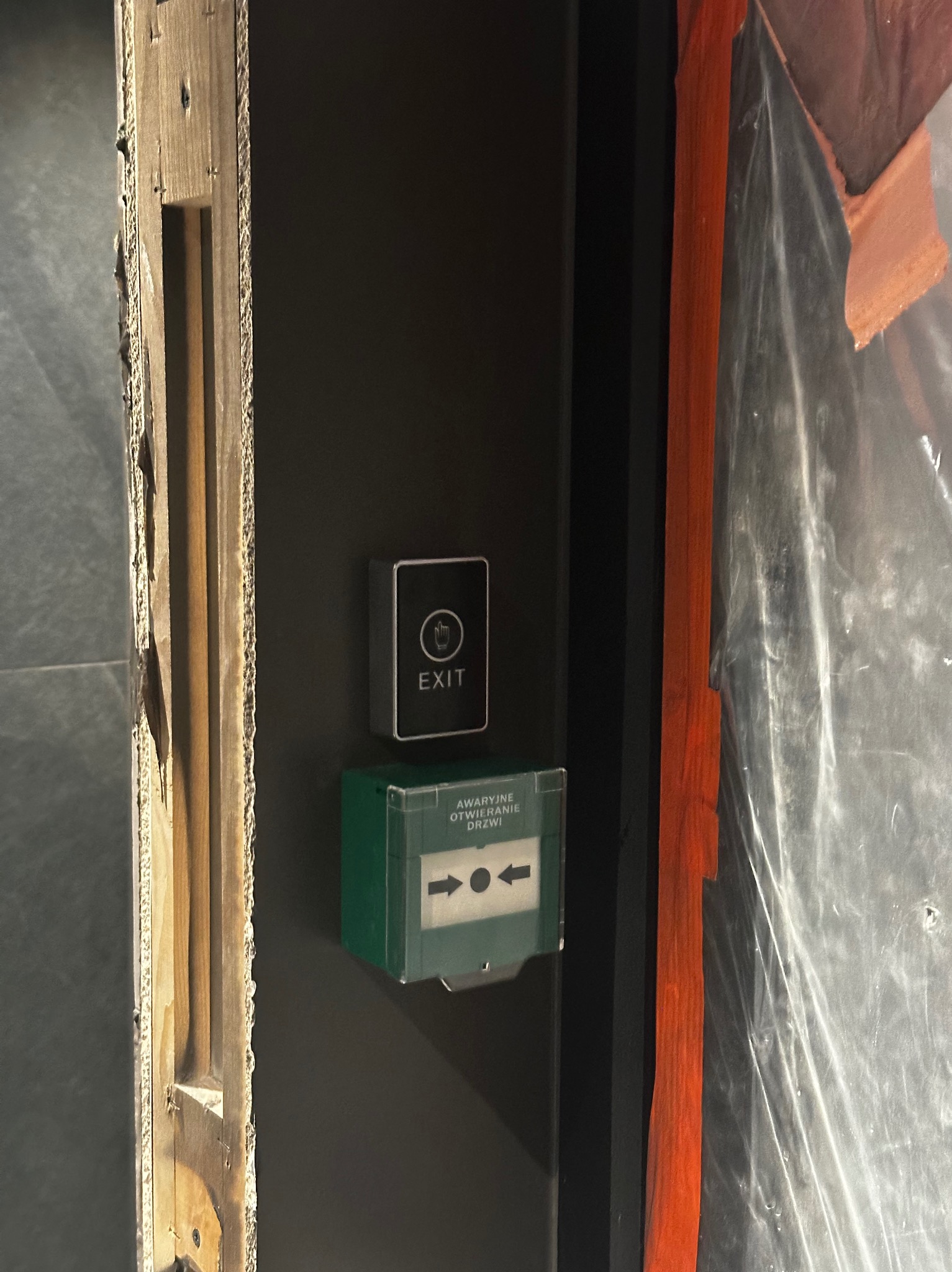 Przycisk wyjścia awaryjnego z zieloną obudową i strzałkami, zamontowany na czarnej ścianie obok drewnianej ościeżnicy i folii ochronnej.