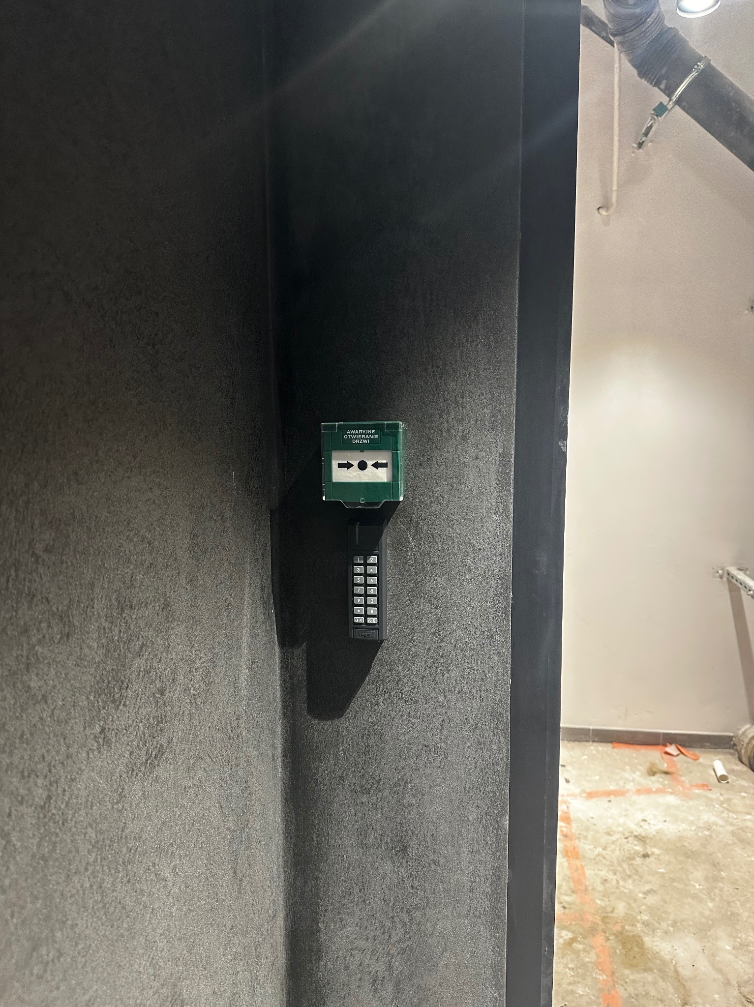 Szary panel z przyciskiem awaryjnego otwierania drzwi i klawiaturą numeryczną, zamontowany na ścianie wewnątrz budynku. Widoczny fragment instalacji pod sufitem.