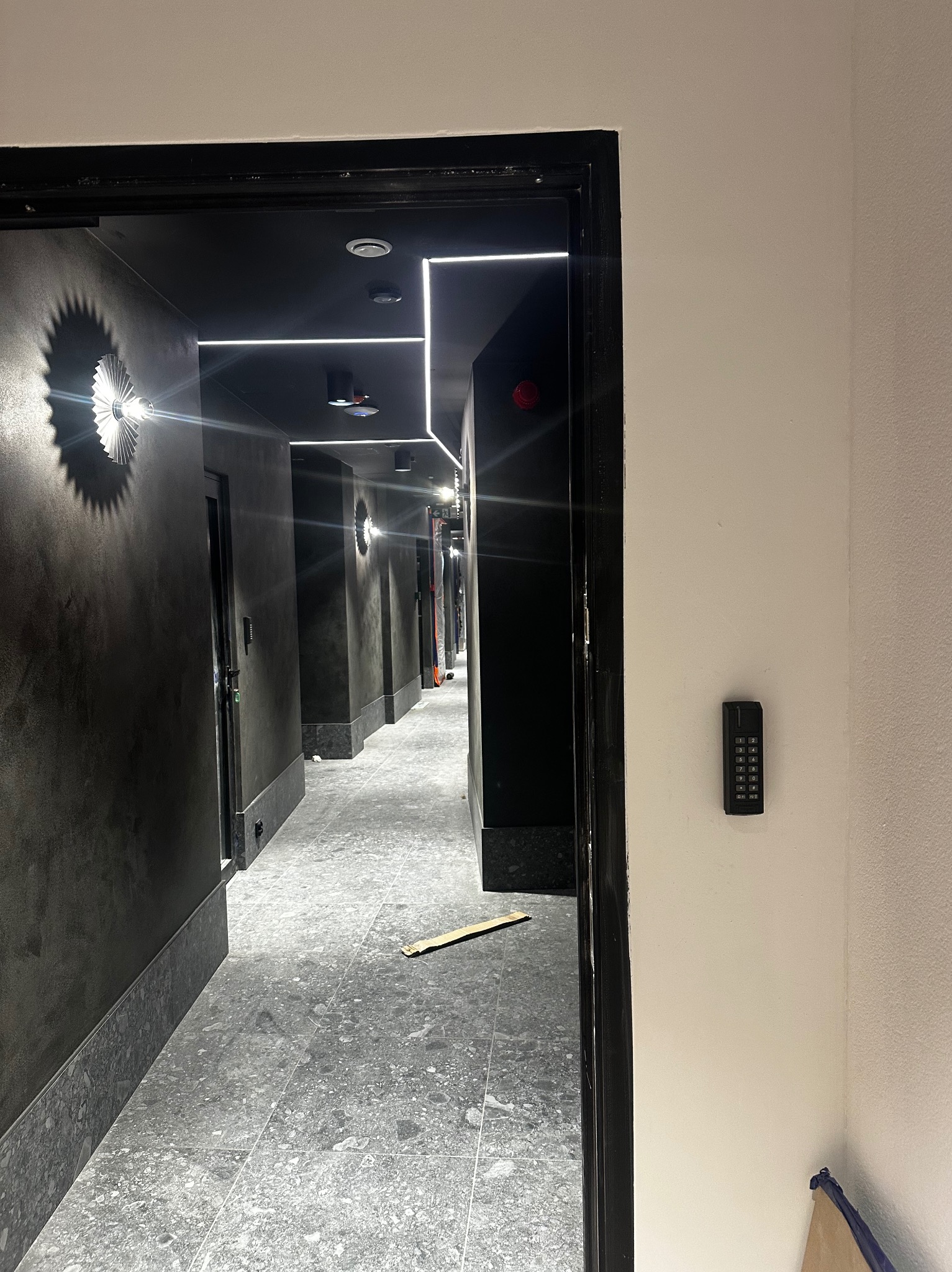Długi korytarz z szarymi ścianami i podłogą, oświetlony liniowymi lampami LED na suficie. Na ścianie widoczny panel kontrolny dostępu z klawiaturą numeryczną.