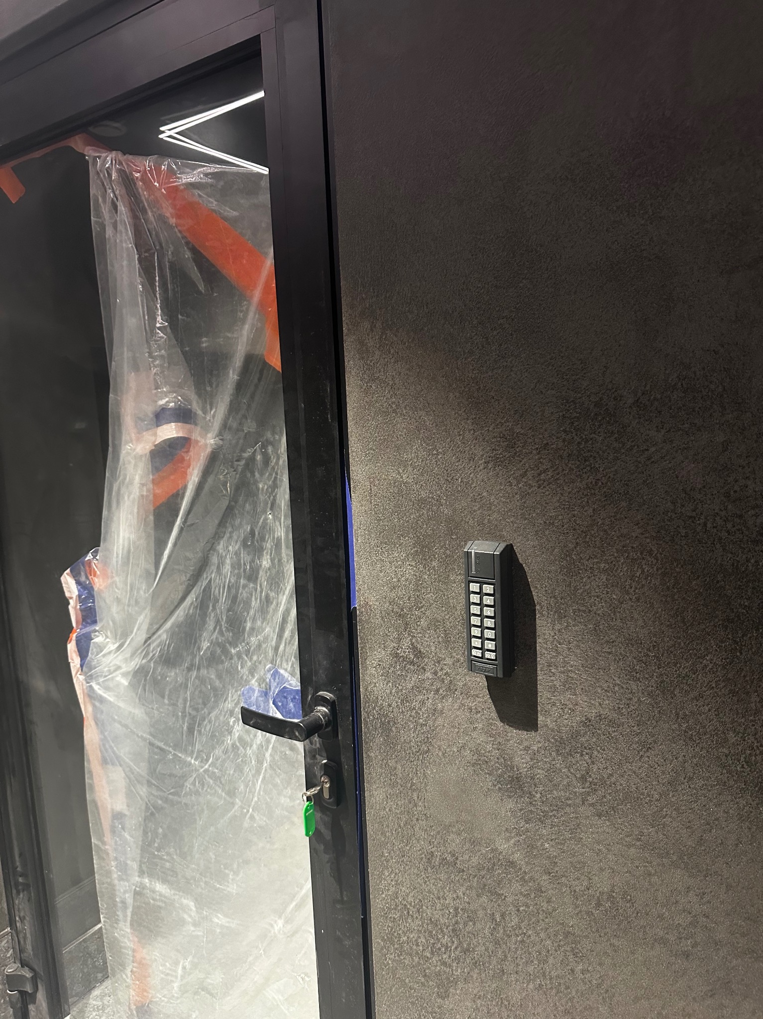 Szary panel ścienny z zamontowanym panelem dostępu kodem numerycznym, widoczny fragment drzwi z folią ochronną, sugerujący montaż systemu kontroli dostępu.