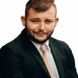 Portret mężczyzny z brodą w czarnym garniturze i jasnym krawacie, uśmiechającego się do kamery na jednolitym tle. Profesjonalny wizerunek, potencjalnie dla firmy.