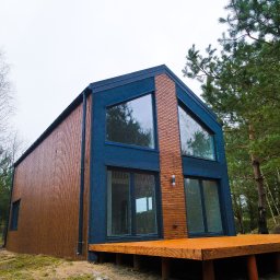 Hocomo Group sp. z o.o. - Nowoczesny dom szkieletowy z drewnianym tarasem i dużymi oknami, otoczony drzewami na tle pochmurnego nieba. Widoczne detale elewacji i konstrukcji.