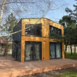Hocomo Group sp. z o.o. - Nowoczesny, dwukondygnacyjny dom szkieletowy z drewnianą elewacją i dużymi oknami, otoczony zielenią i drewnianym tarasem. Widoczna antena satelitarna.