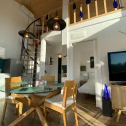 Jasne wnętrze domu szkieletowego z drewnianą spiralną klatką schodową, szklanym stołem i widokiem na sypialnię. Elementy dekoracyjne z butelek.