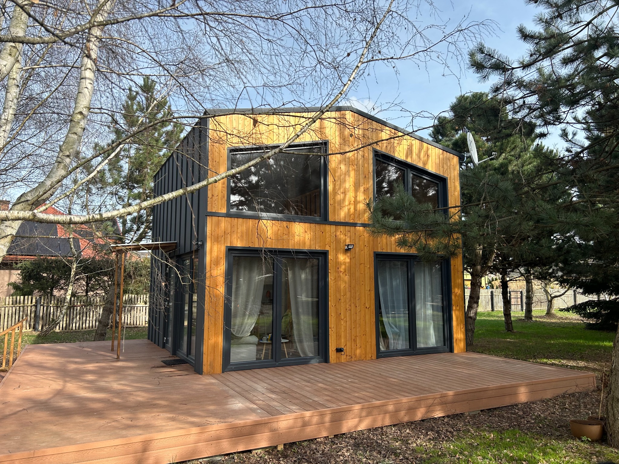 Nowoczesny, dwukondygnacyjny dom szkieletowy z drewnianą elewacją i dużymi oknami, otoczony zielenią i drewnianym tarasem. Widoczna antena satelitarna.