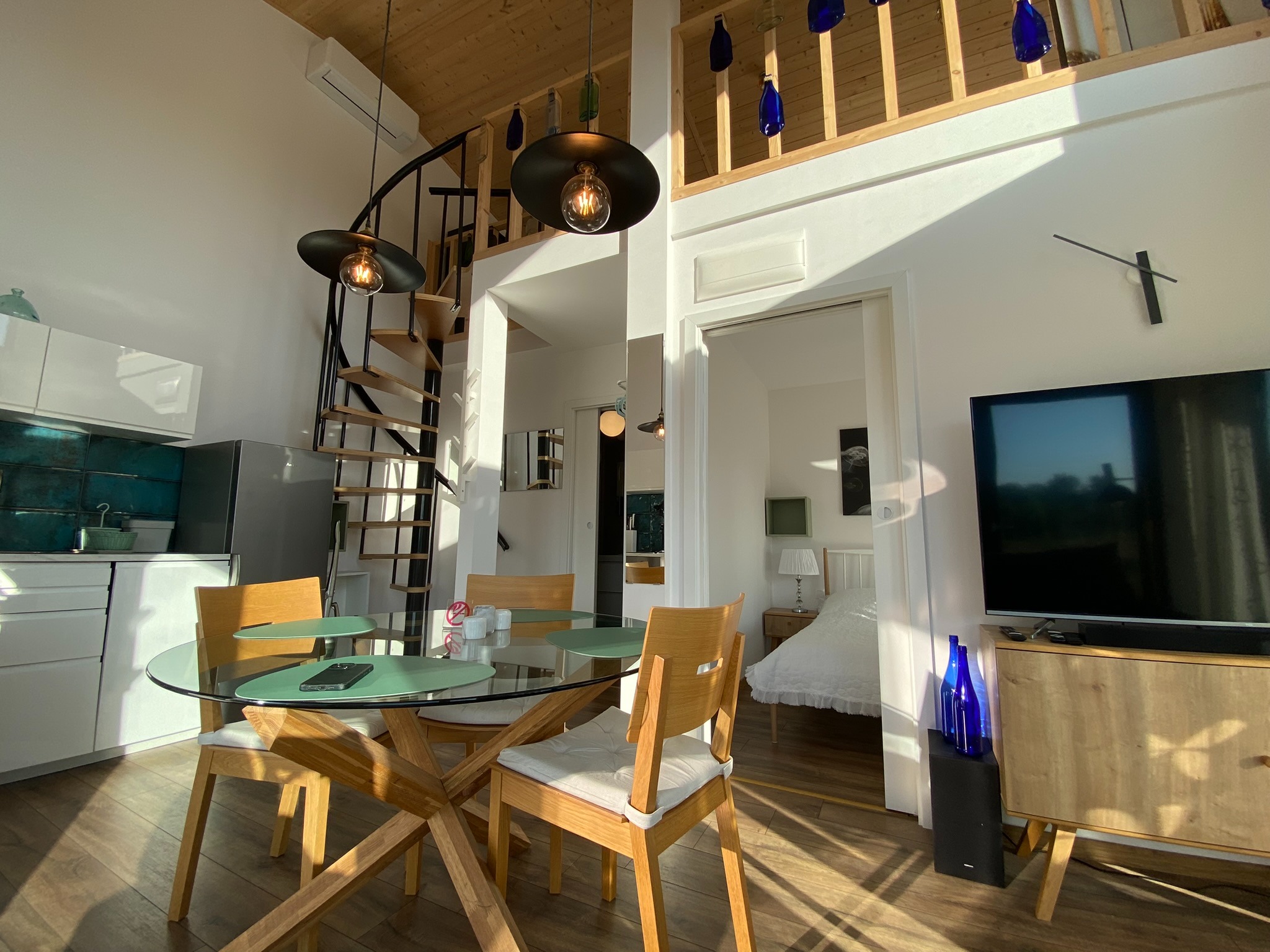 Jasne wnętrze domu szkieletowego z drewnianą spiralną klatką schodową, szklanym stołem i widokiem na sypialnię. Elementy dekoracyjne z butelek.