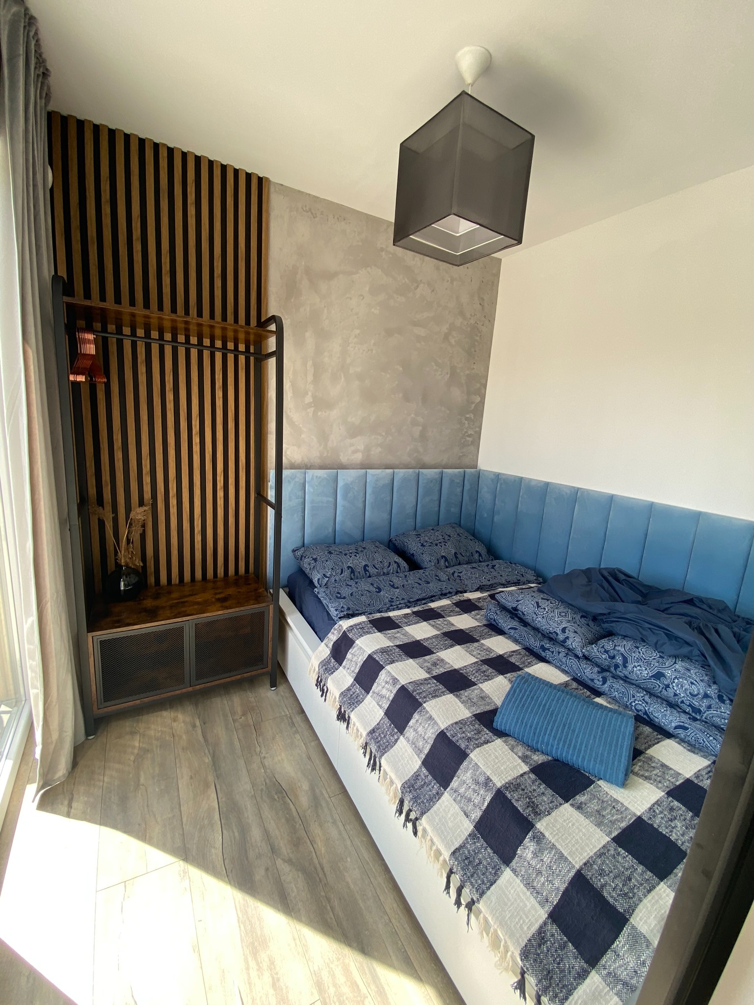 Jasna sypialnia z łóżkiem w niebieskiej tonacji, drewnianą ścianką działową i metalowym regałem. Parkiet w odcieniach szarości. Nowoczesne wnętrze.