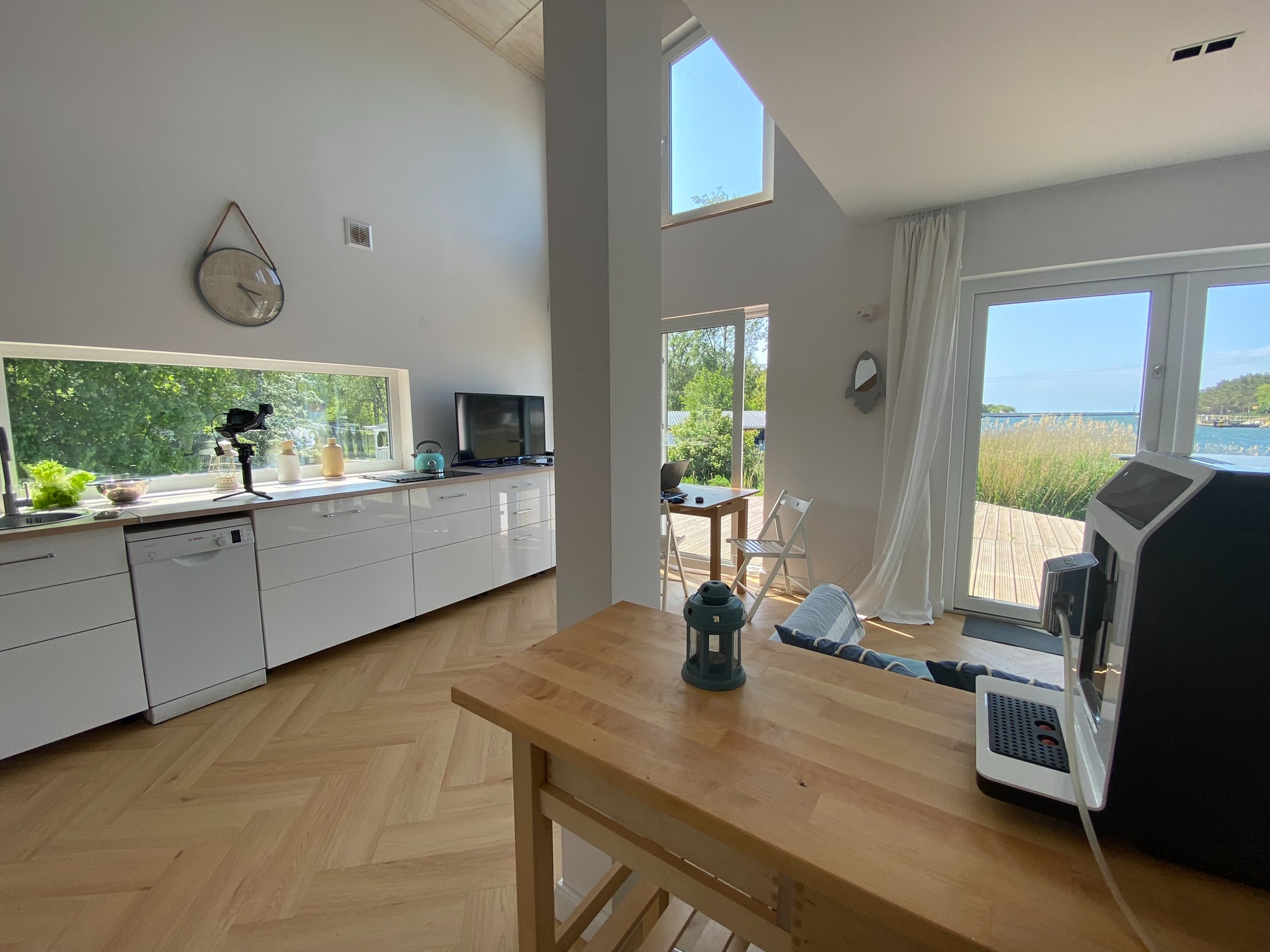 Jasna, nowoczesna kuchnia z widokiem na zewnątrz. Parkiet w jodełkę, białe meble kuchenne i duże okno z widokiem na zieleń i wodę. Minimalistyczny zegar na ścianie.