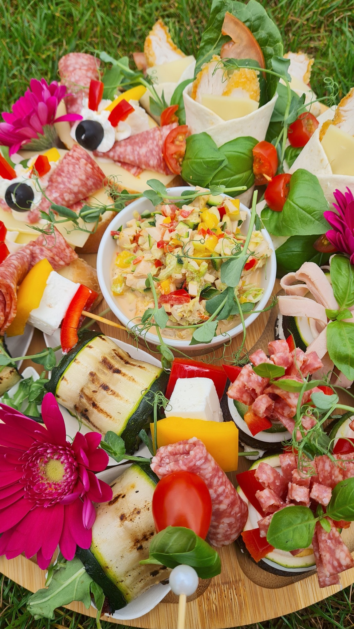 Bogato zdobiona deska przekąsek na trawie: grillowana cukinia, sałatka warzywna, szaszłyki z serem i papryką, salami, ser i kwiaty gerbery tworzą apetyczną kompozycję.