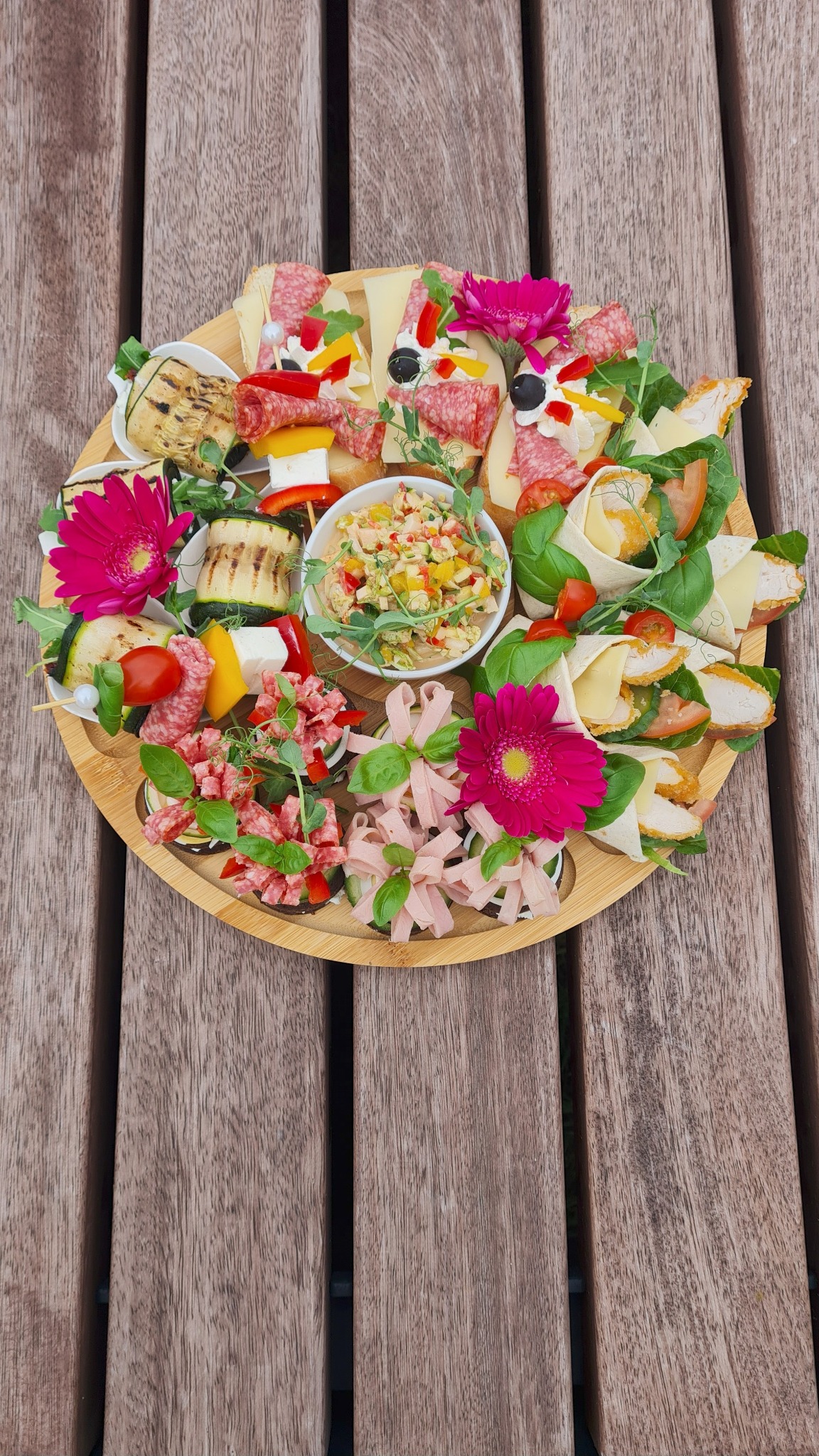 Dekoracyjna deska z przekąskami: roladki z kurczakiem, koreczki z salami i warzywami, sałatka. Kompozycja ozdobiona różowymi kwiatami na drewnianym stole.