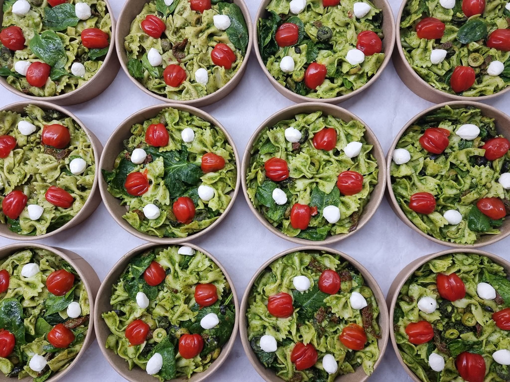 Dwanaście porcji sałatki farfalle z pesto, pomidorkami cherry, mozzarellą i szpinakiem, podane w ekologicznych, papierowych miseczkach.