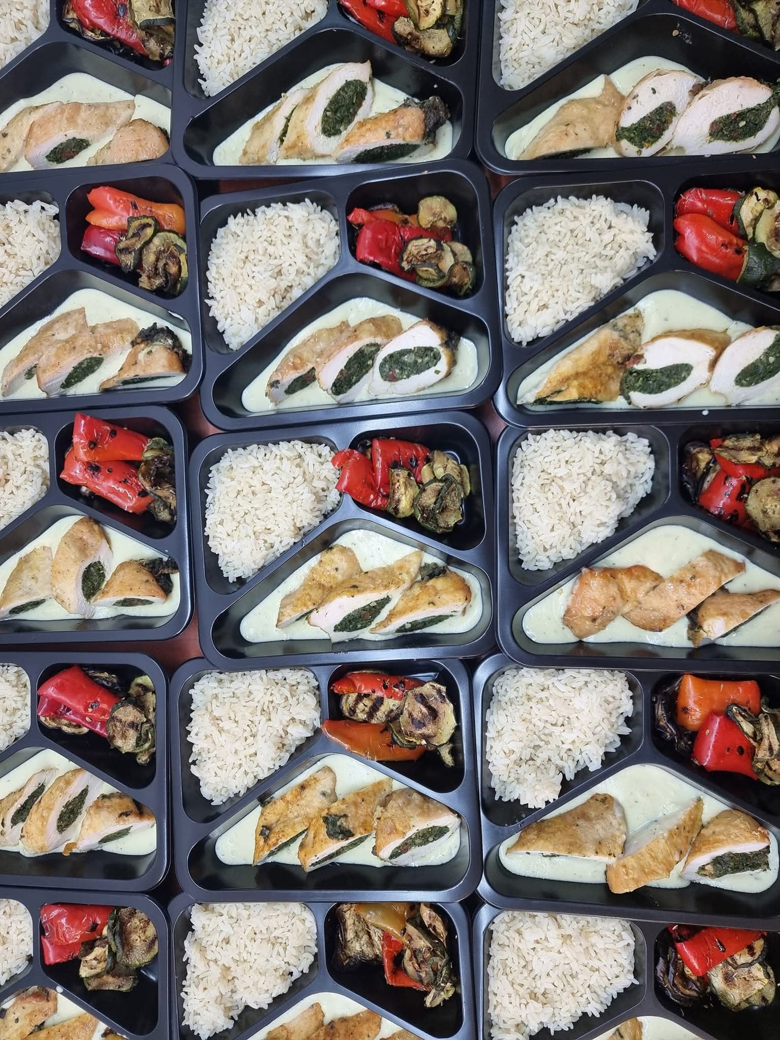 Zestaw dań w czarnych pojemnikach: roladki z kurczaka ze szpinakiem w sosie, ryż i grillowane warzywa (papryka, cukinia). Widok z góry na cateringowe porcje.