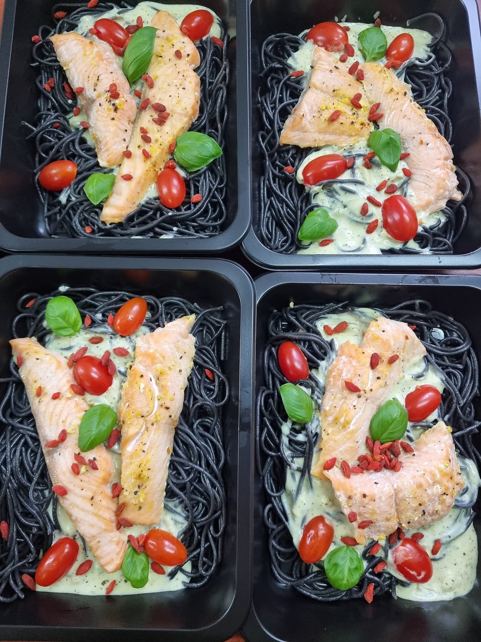 Cztery porcje czarnego makaronu z łososiem, sosem, pomidorkami i bazylią, podane w czarnych pojemnikach. Danie gotowe do spożycia, catering dietetyczny.