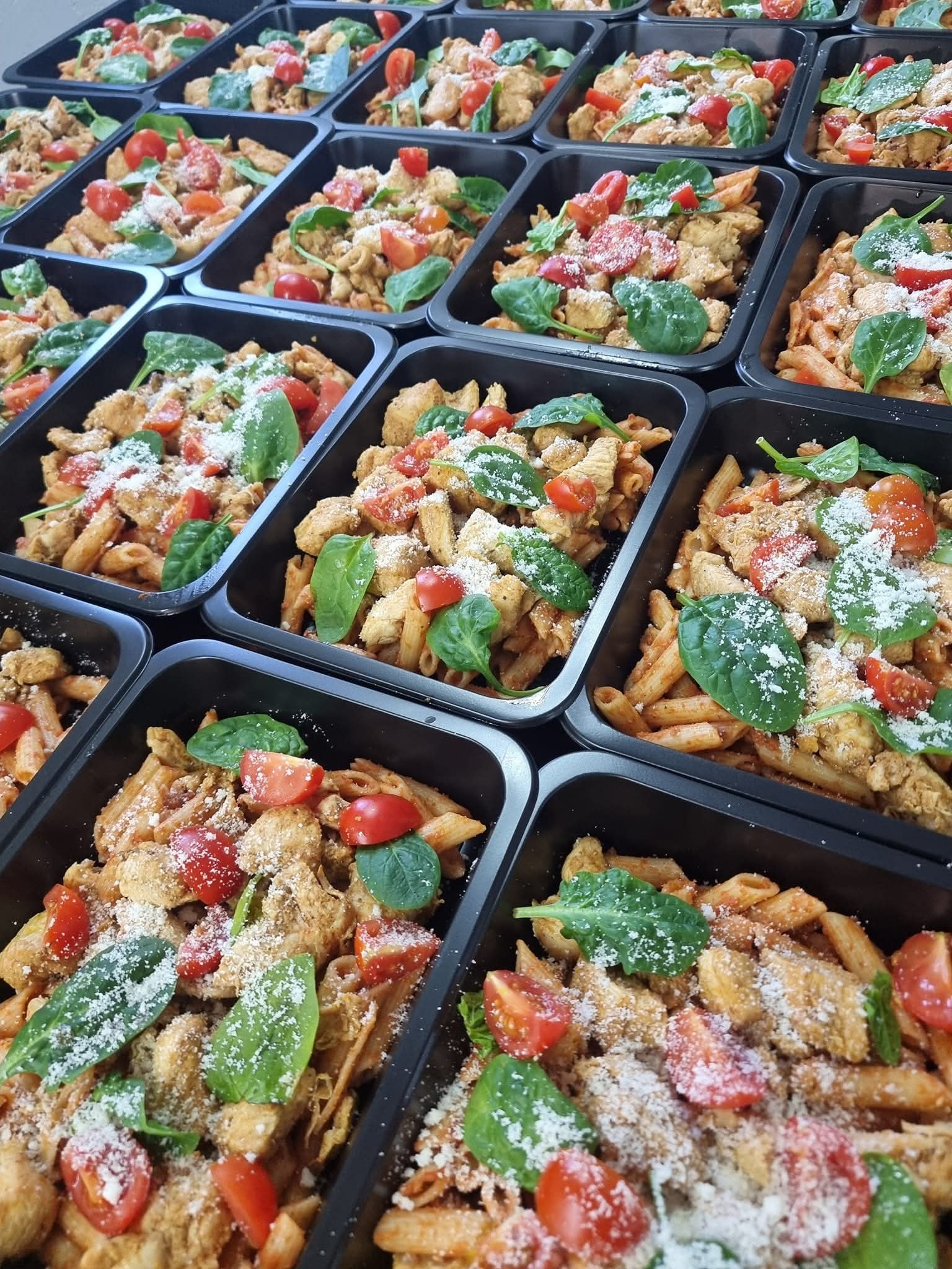 Penne z kurczakiem, pomidorami, szpinakiem i parmezanem w czarnych pojemnikach. Gotowe porcje cateringu dietetycznego ułożone rzędami.