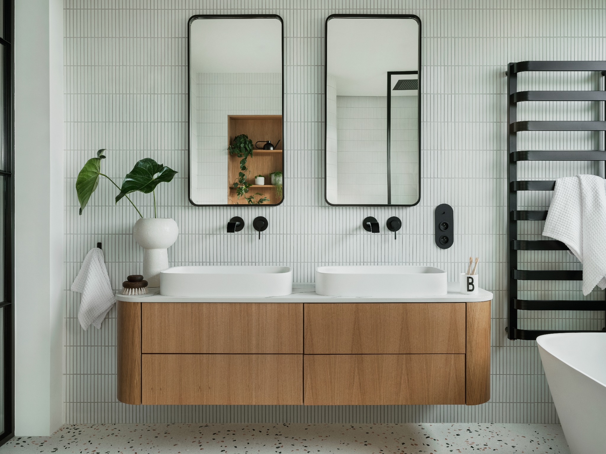 Nowoczesna łazienka z drewnianą szafką pod umywalkami, dwoma lustrami w czarnych ramach i grzejnikiem drabinkowym. Minimalistyczny design, jasne płytki i terrazzo na podłodze.