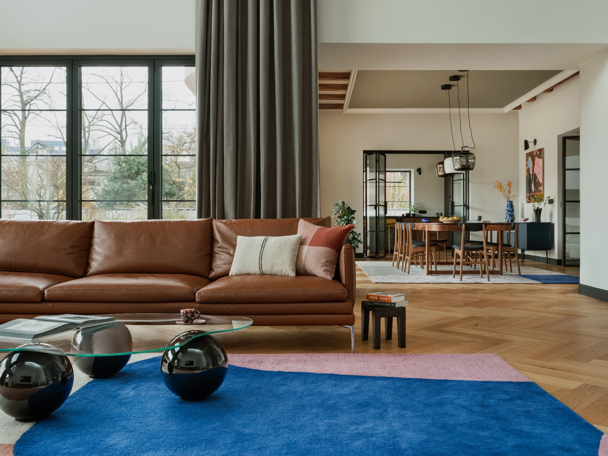 Stylowy salon z brązową skórzaną sofą, geometrycznym dywanem i szklanym stolikiem kawowym. Widok na jadalnię przez przeszklone drzwi.