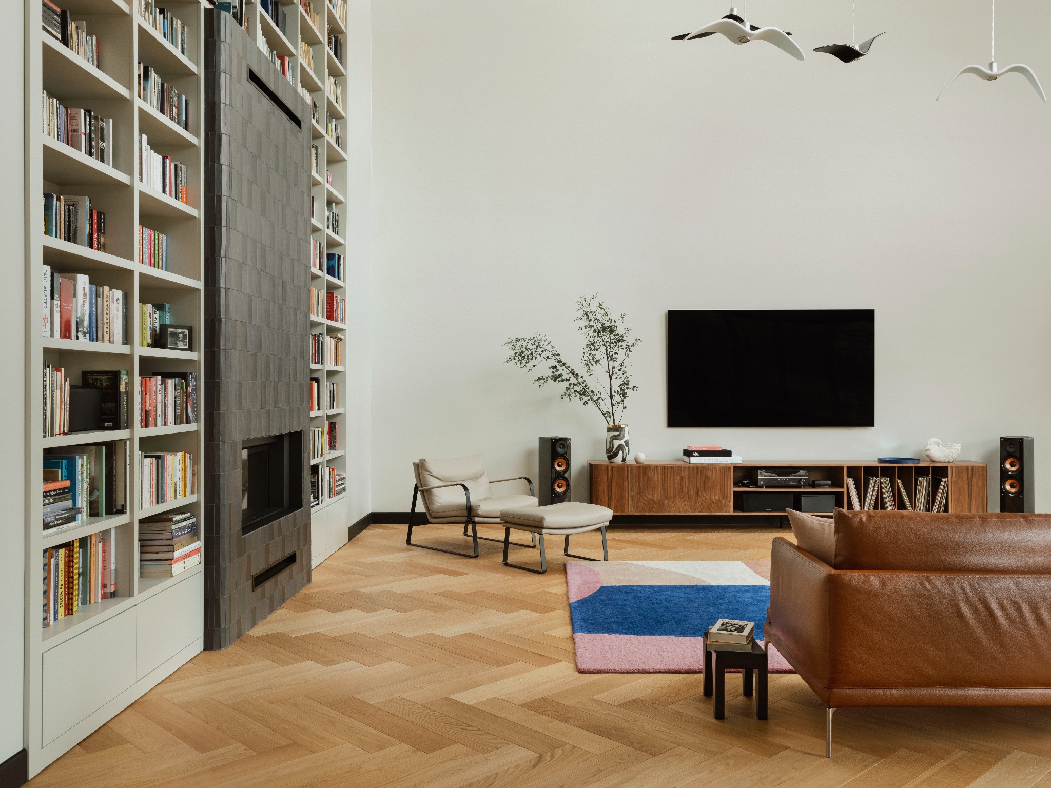 Elegancki salon z wysokim regałem na książki, kominkiem z szarej cegły, drewnianą podłogą w jodełkę i designerskimi lampami w kształcie ptaków.