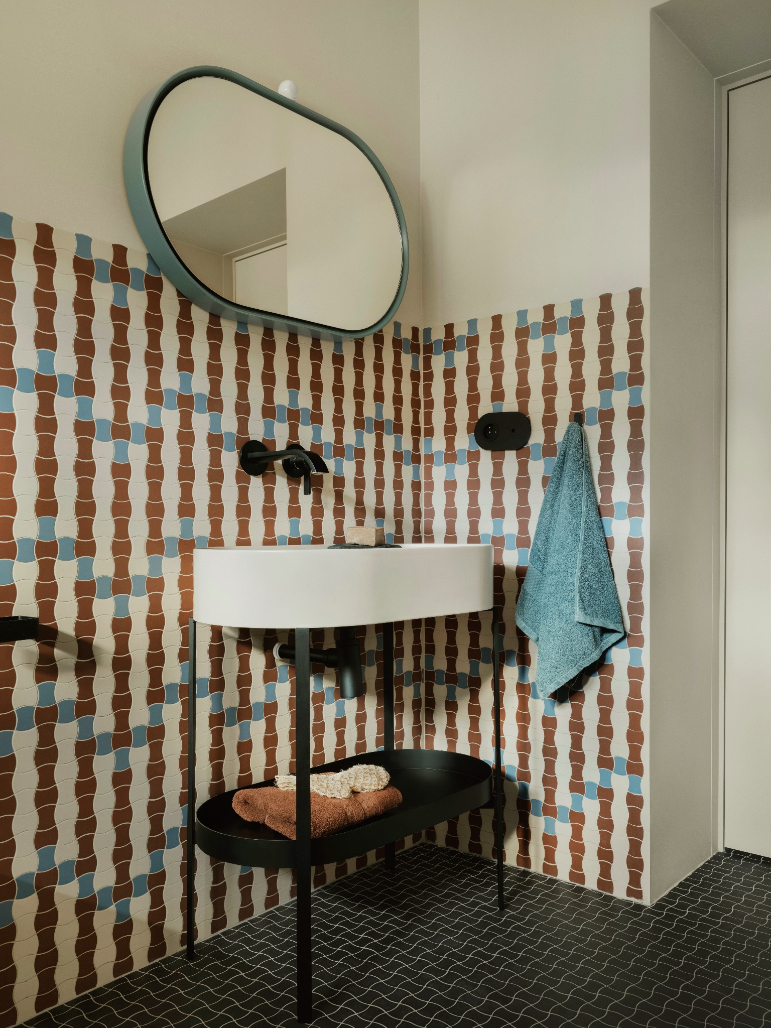 Nowoczesna łazienka z umywalką na czarnym stelażu, owalnym lustrem w turkusowej ramie i wzorzystymi płytkami w odcieniach brązu i błękitu. Minimalistyczny design.