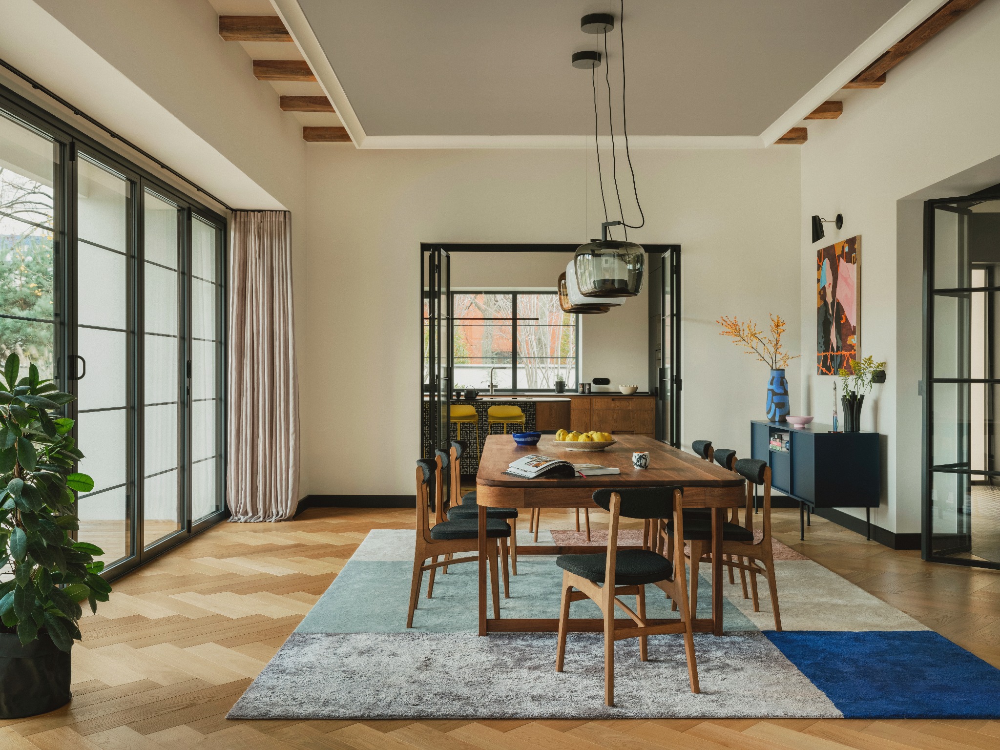 Jasna jadalnia z drewnianym stołem i krzesłami na patchworkowym dywanie. W tle widoczna kuchnia i salon, parkiet w jodełkę, duże okna z czarną ramą.