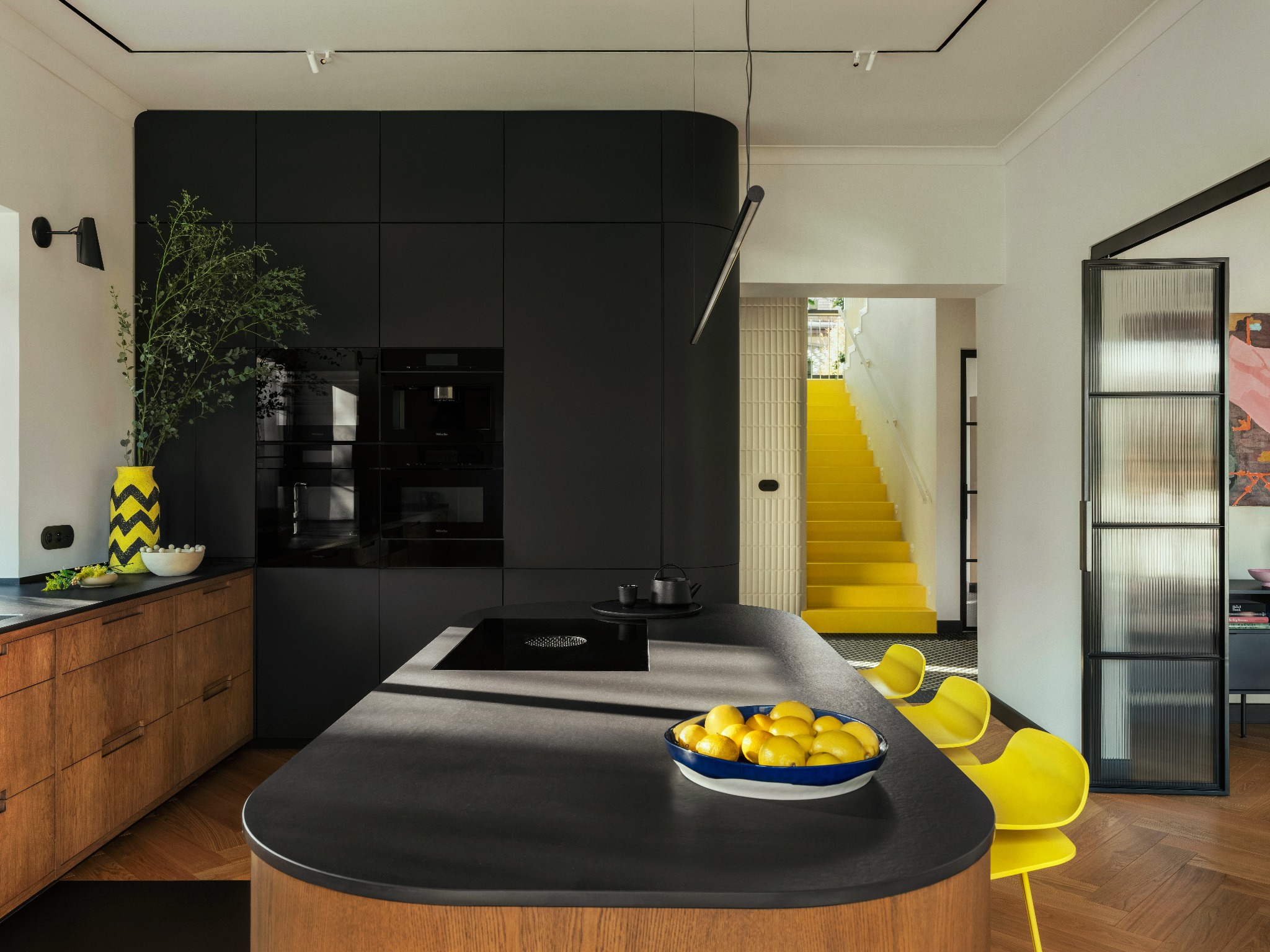 Nowoczesna kuchnia w Łodzi z czarnymi szafkami, drewnianym blatem i żółtymi akcentami, w tym schodami i krzesłami. Widok na przeszklone drzwi i dekoracje.
