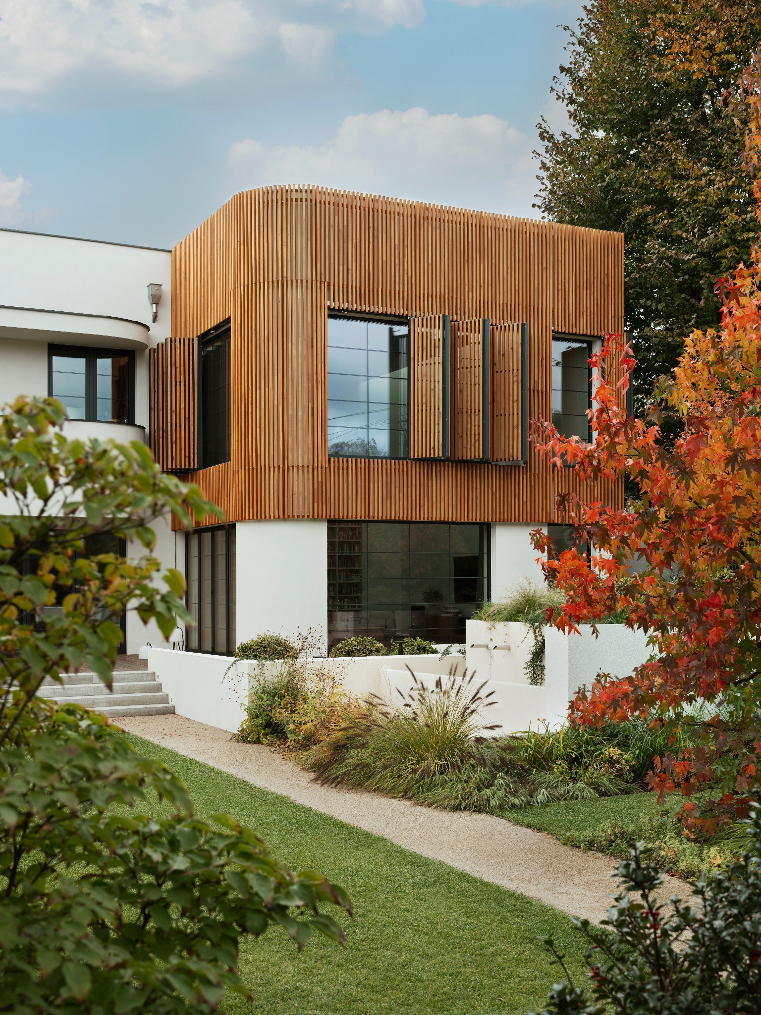 Nowoczesna elewacja domu z drewnianymi lamelami i otwartymi okiennicami, otoczona zielenią. Widok z perspektywy ogrodu, jesienne barwy drzew.