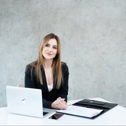 Concept Pro. Żaneta Gasek - Profesjonalna agentka nieruchomości siedzi przy biurku z laptopem i dokumentami. Uśmiechnięta kobieta w czarnym żakiecie, jasne tło. Gotowa do pracy.