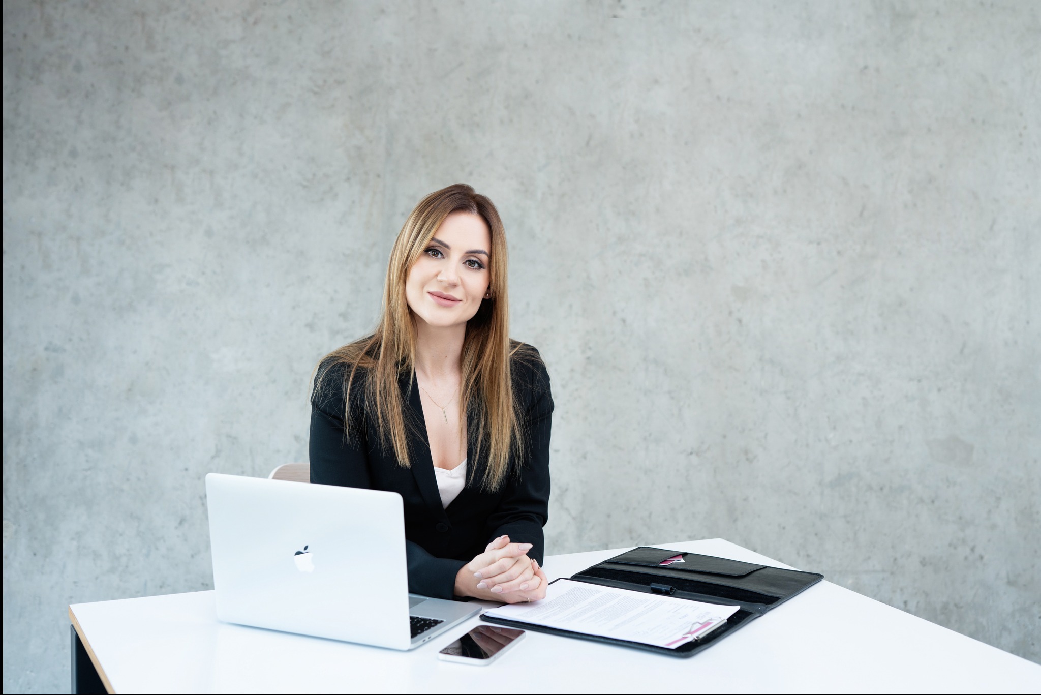 Profesjonalna agentka nieruchomości siedzi przy biurku z laptopem i dokumentami. Uśmiechnięta kobieta w czarnym żakiecie, jasne tło. Gotowa do pracy.