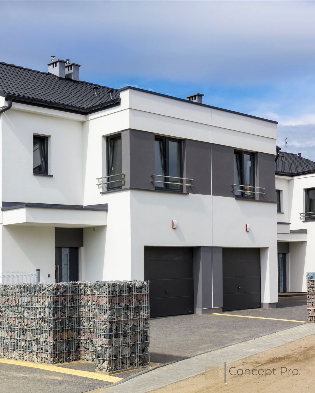 Nowoczesny, dwukondygnacyjny dom w odcieniach bieli i szarości z czarną bramą garażową. Gabionowe ogrodzenie z kamienia w tle.