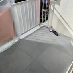 Wyłożony szarymi płytkami balkon z osłoną z pleksi i zabezpieczoną folią częścią. Widoczny mop do podłogi. Słoneczny dzień.