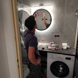 Dariusz Ragan - Montaż okrągłego lustra z podświetleniem LED w nowoczesnej łazience z szarymi płytkami. Widoczne narzędzia i pralka Electrolux, etap prac wykończeniowych.