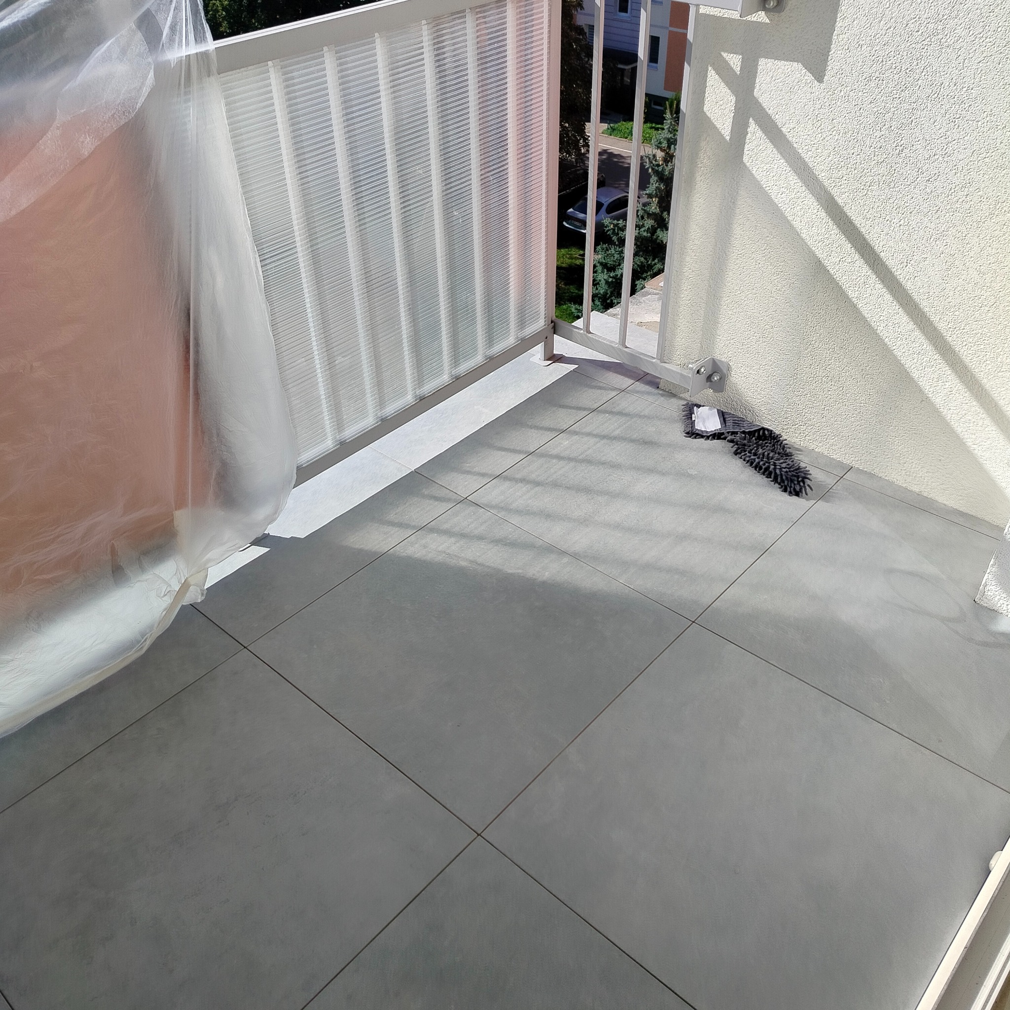 Wyłożony szarymi płytkami balkon z osłoną z pleksi i zabezpieczoną folią częścią. Widoczny mop do podłogi. Słoneczny dzień.