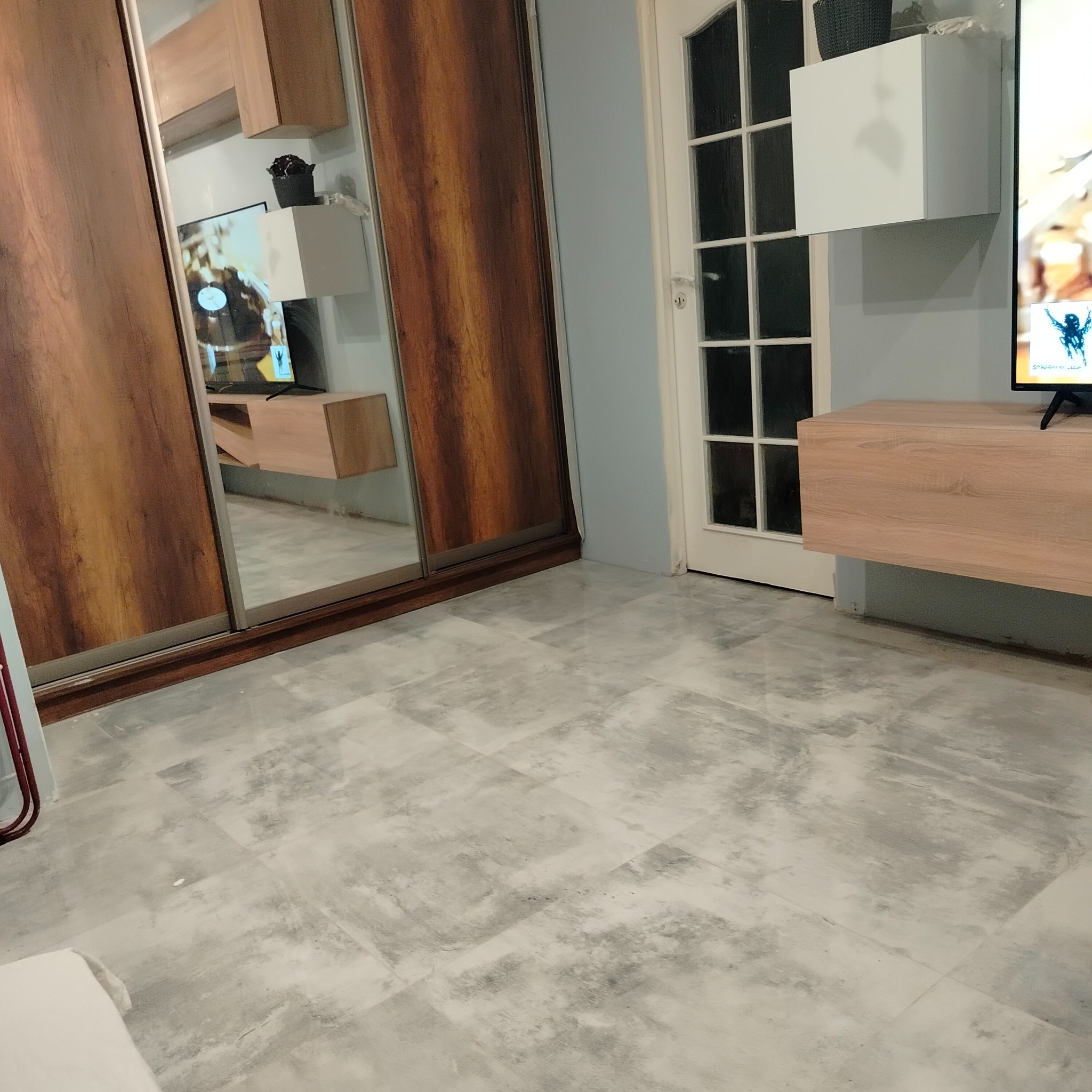 Nowoczesny salon z szarą podłogą z płytek, przesuwną szafą z lustrem odbijającym telewizor i szafki RTV. Minimalistyczny design.