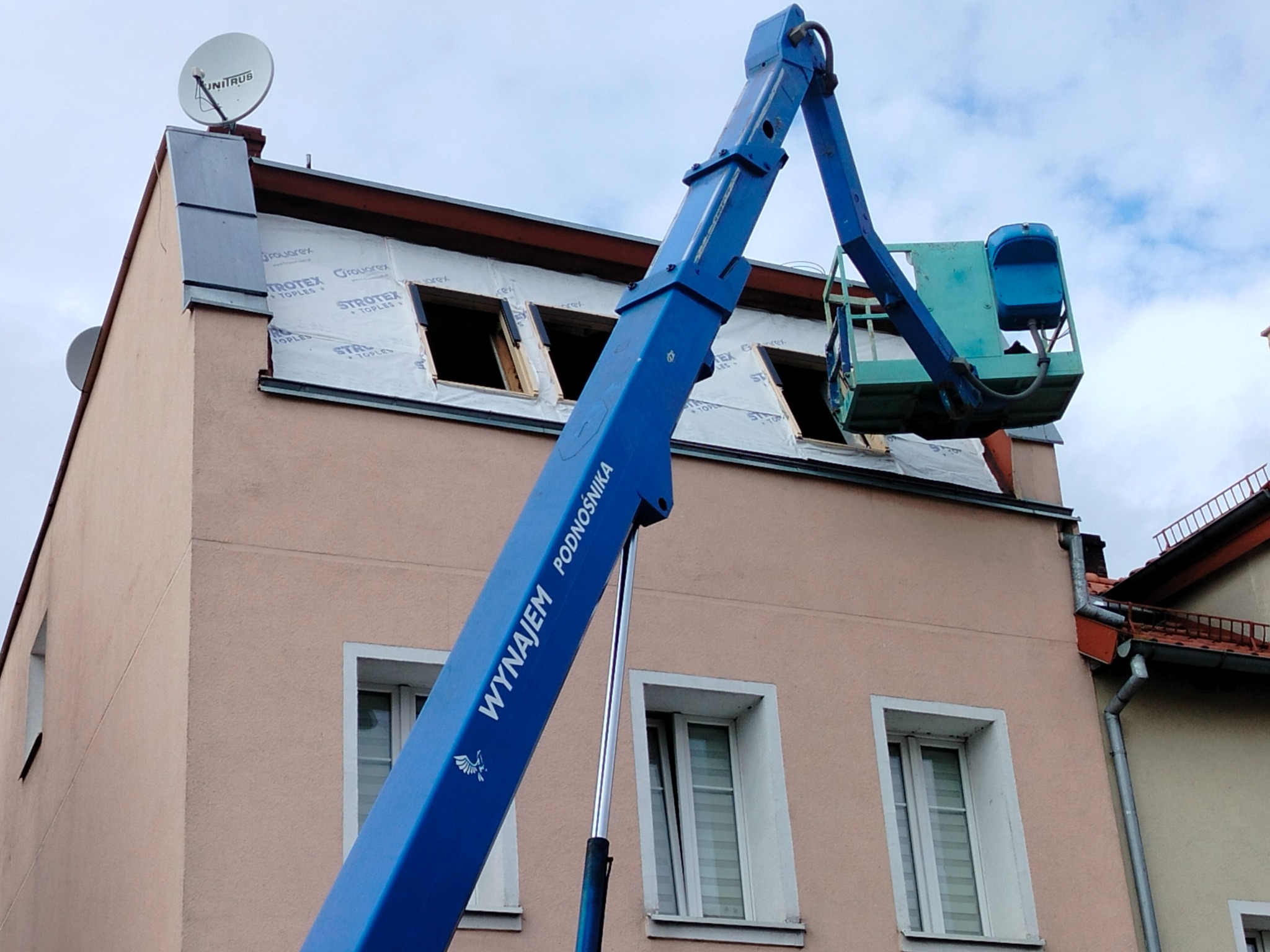 Podnośnik koszowy na tle remontowanej kamienicy z oknami zabezpieczonymi folią. Widoczna antena satelitarna na dachu i fragment sąsiedniego budynku.