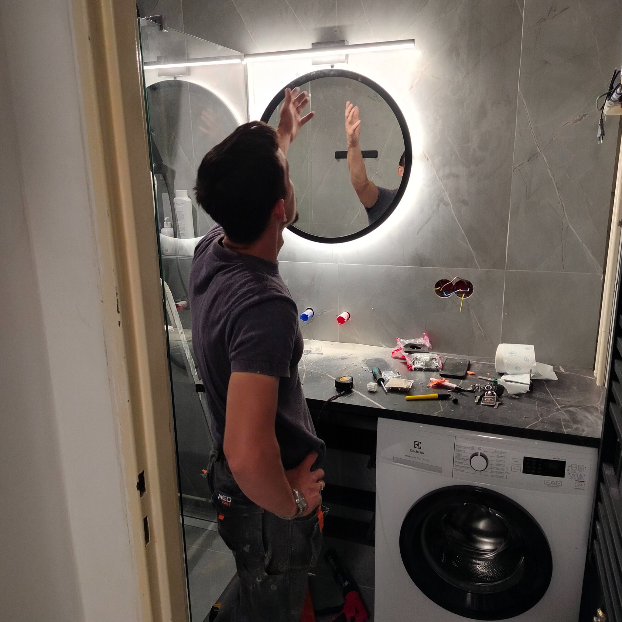 Montaż okrągłego lustra z podświetleniem LED w nowoczesnej łazience z szarymi płytkami. Widoczne narzędzia i pralka Electrolux, etap prac wykończeniowych.