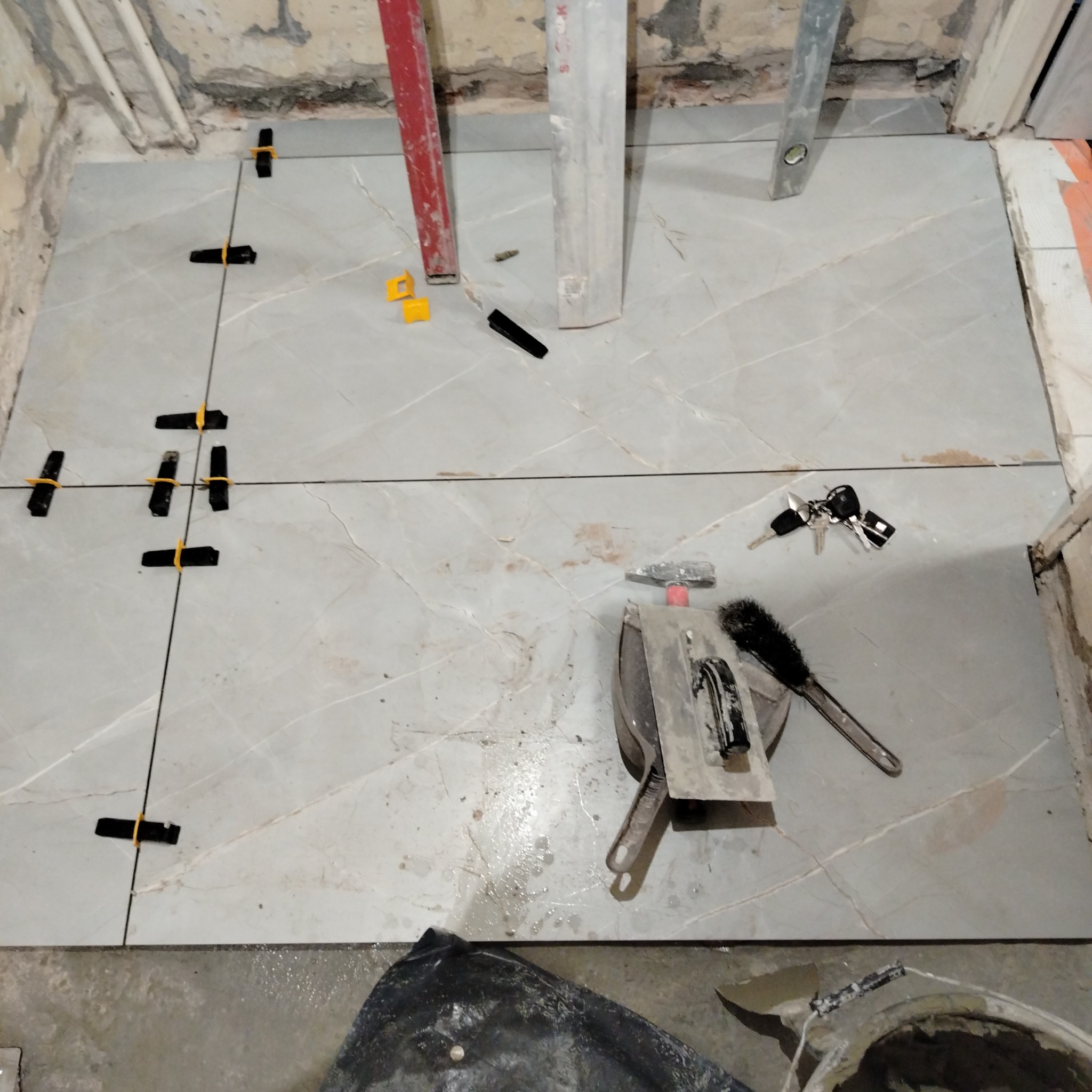 Układanie szarych płytek z marmurowym wzorem, z użyciem poziomnic i klipsów poziomujących. Widoczne narzędzia glazurnicze i wiązka kluczy.