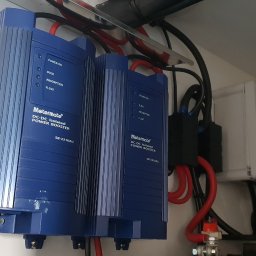 Magazyn Energii - Inteligentne Domy - Elektryka - VoltLogic - Dwa niebieskie izolowane wzmacniacze mocy DC-DC Metermate SB-2210AU z okablowaniem wewnątrz białej szafki. Widoczne diody LED, złącza i przewody.