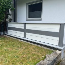 Finezja Katarzyna Olejarz - Nowoczesne ogrodzenie panelowe w odcieniach szarości i bieli, z widoczną strukturą drewna, zamontowane przy budynku z zielonym trawnikiem. Ujęcie z poziomu gruntu.