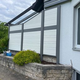 Finezja Katarzyna Olejarz - Nowoczesny, biało-szary panel ogrodzeniowy z poziomymi deskami, osadzony na kamiennym murku rabaty z zielenią. Widok z boku, fragment domu z oknem w tle.