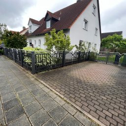 Finezja Katarzyna Olejarz - Nowoczesne, grafitowe ogrodzenie posesji z ozdobnymi elementami, otaczające dom z czerwonym dachem. Widok z chodnika z kostki brukowej na podjazd.