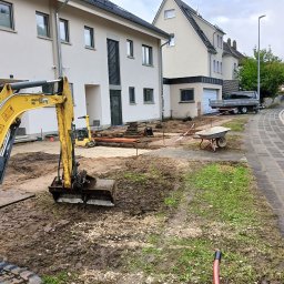 Finezja Katarzyna Olejarz - Żółta mini koparka na placu budowy przed domem. Widoczne wykopane fundamenty, rury i taczka. Etap przygotowania terenu pod budowę lub prace ogrodowe.