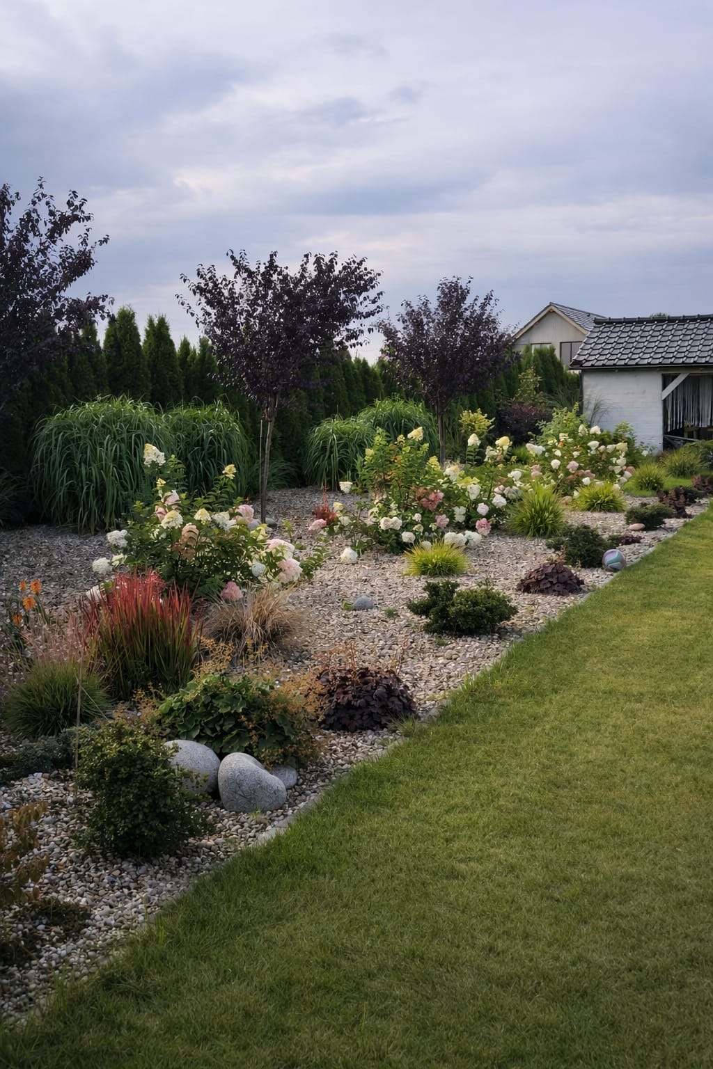 Ogród w Snowidzy z kwitnącymi hortensjami, ozdobnymi trawami i drzewami o purpurowych liściach na tle zielonych tuj i białego domku.