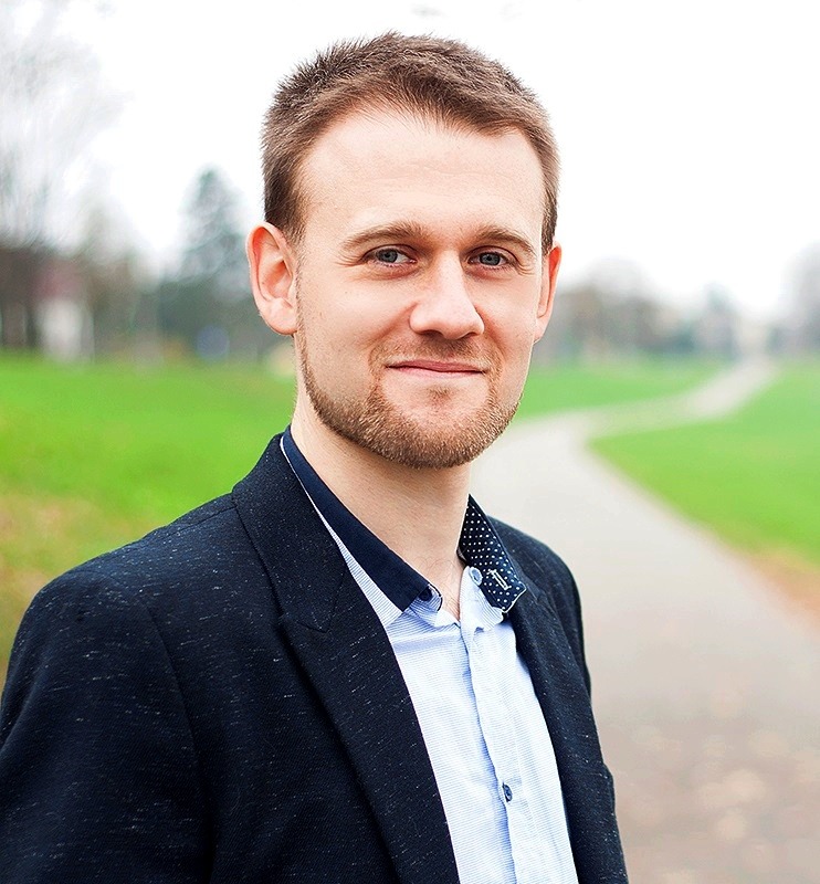 Portret uśmiechniętego mężczyzny w marynarce na tle zielonego parku. Ujęcie z lekkiego dołu, podkreślające profesjonalny wygląd.