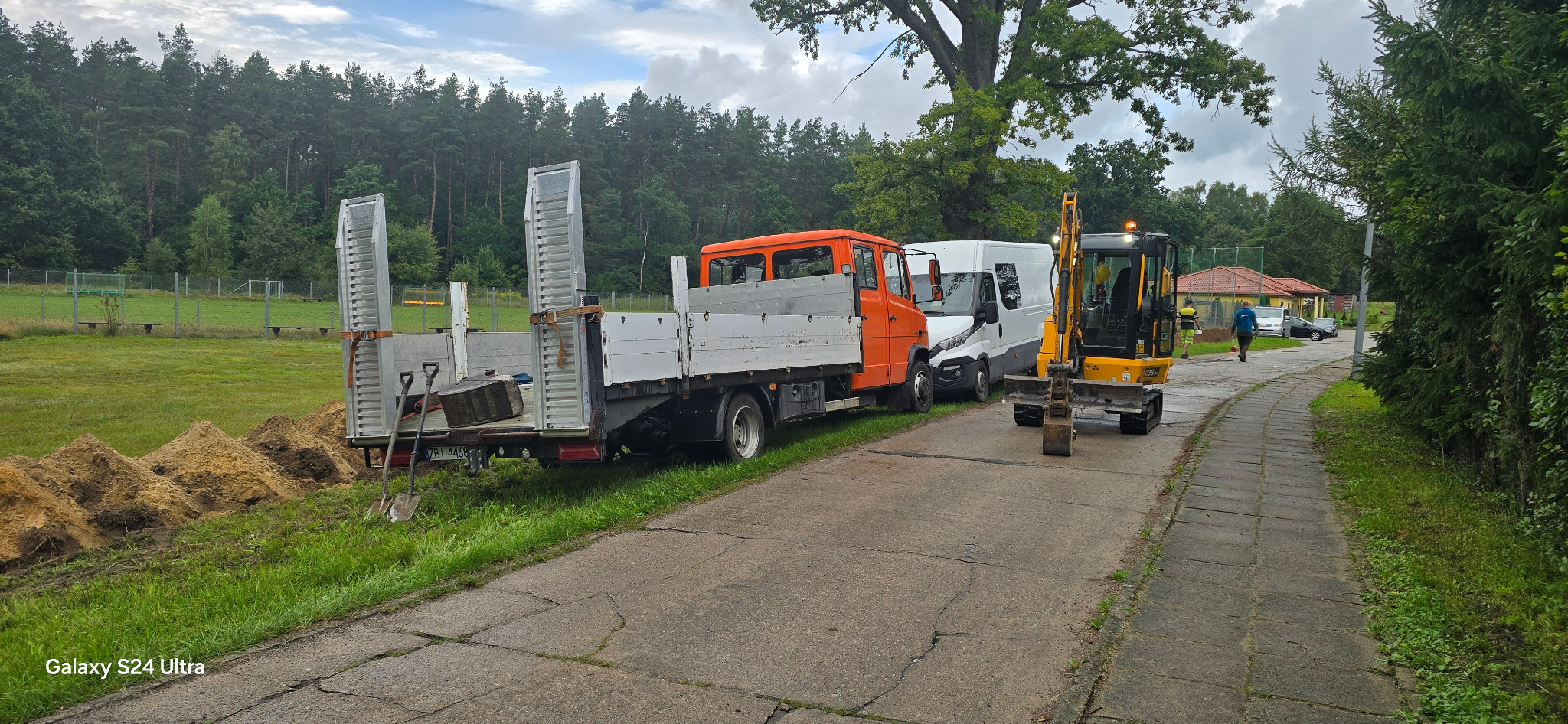 Żółta mini koparka, pomarańczowa ciężarówka i biały bus zaparkowane obok siebie na drodze, za nimi hałda ziemi i teren zielony z drzewami.