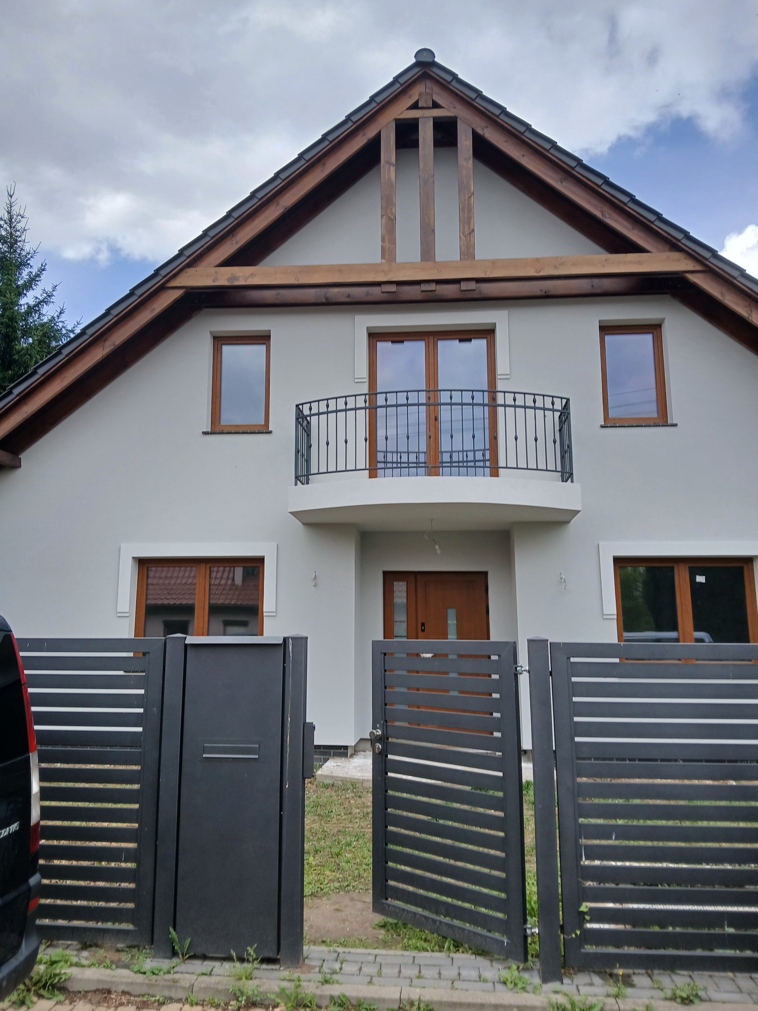 Elewacja domu jednorodzinnego z balkonem i drewnianymi oknami, widoczna przez nowoczesne, poziome ogrodzenie. Szara kolorystyka domu i ciemne ogrodzenie.