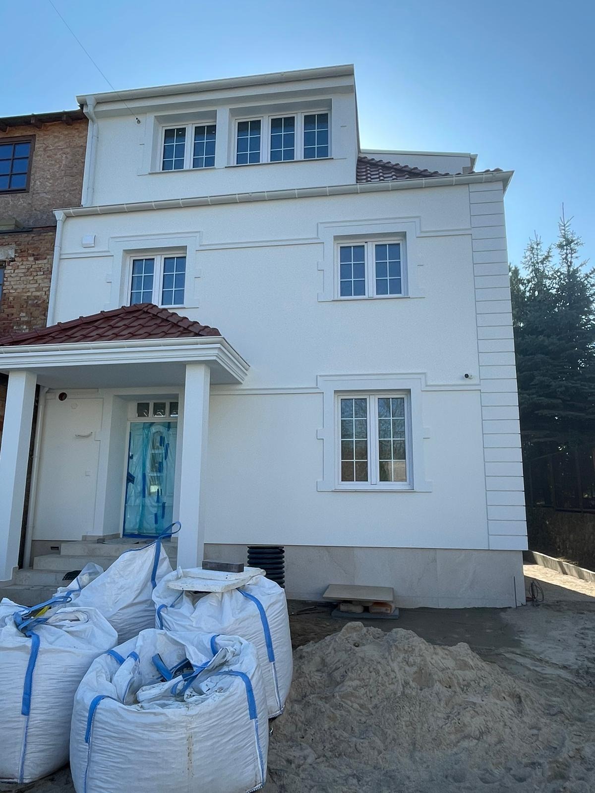 Biała elewacja domu z dekoracyjnymi detalami, worki z materiałami budowlanymi na pierwszym planie. Widoczne okna z białymi ramami i fragment dachu z czerwoną dachówką.