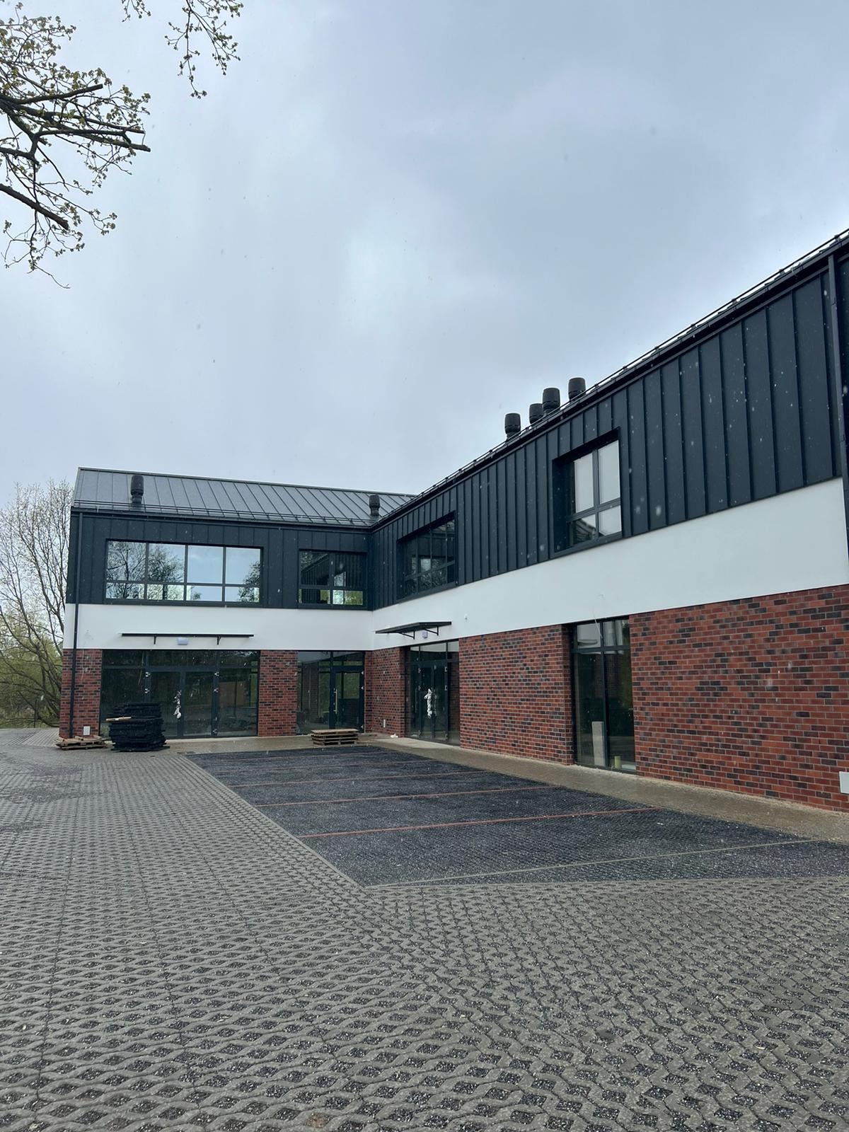 Nowoczesny budynek z elewacją z czerwonej cegły, białego tynku i czarnej blachy trapezowej. Widoczne okna i drzwi. Na pierwszym planie kostka brukowa.