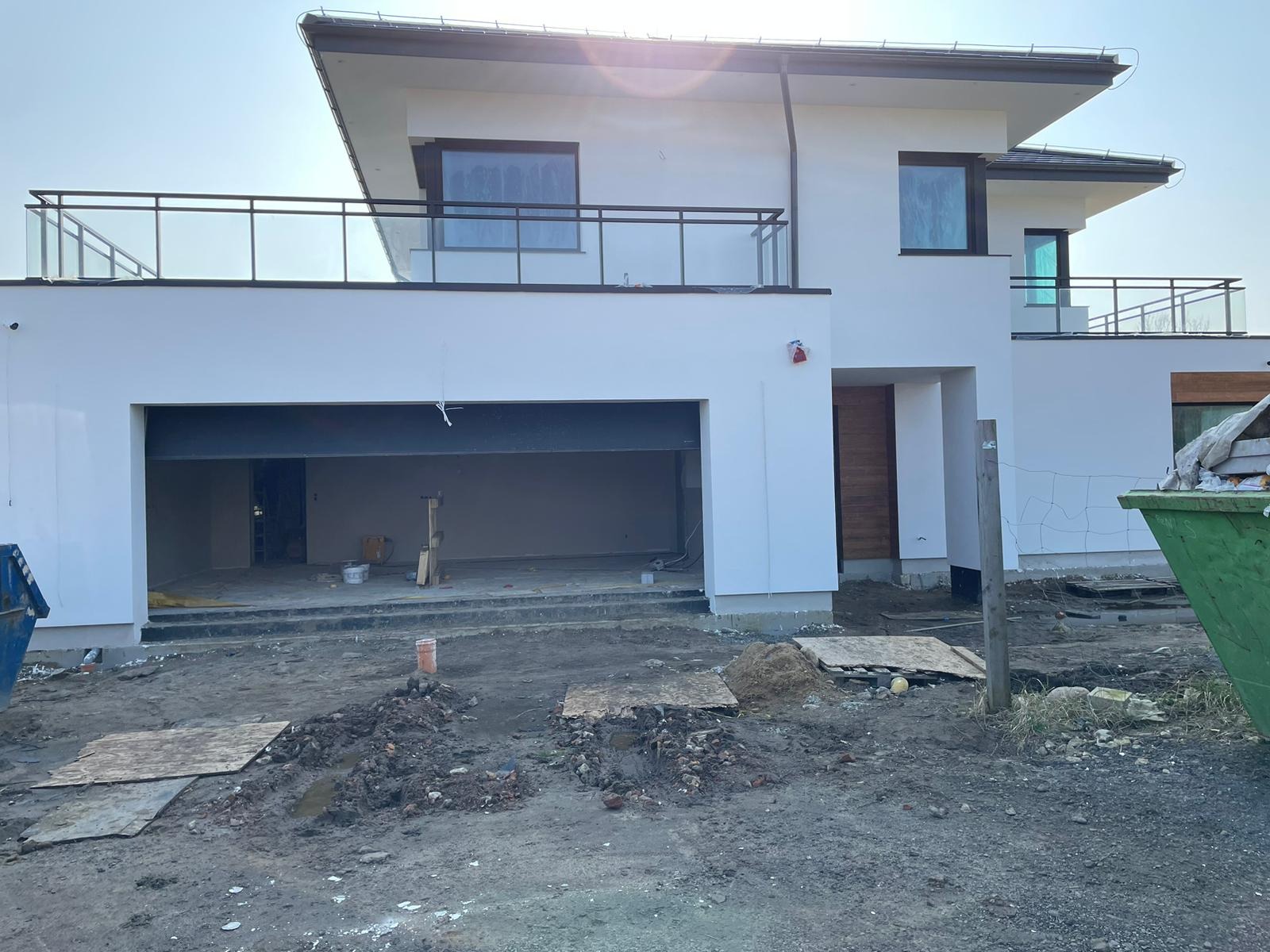 Biały, dwukondygnacyjny dom z balkonami w trakcie budowy. Widoczny otwarty garaż, teren wokół w stanie surowym, kontener na odpady. Słoneczny dzień.