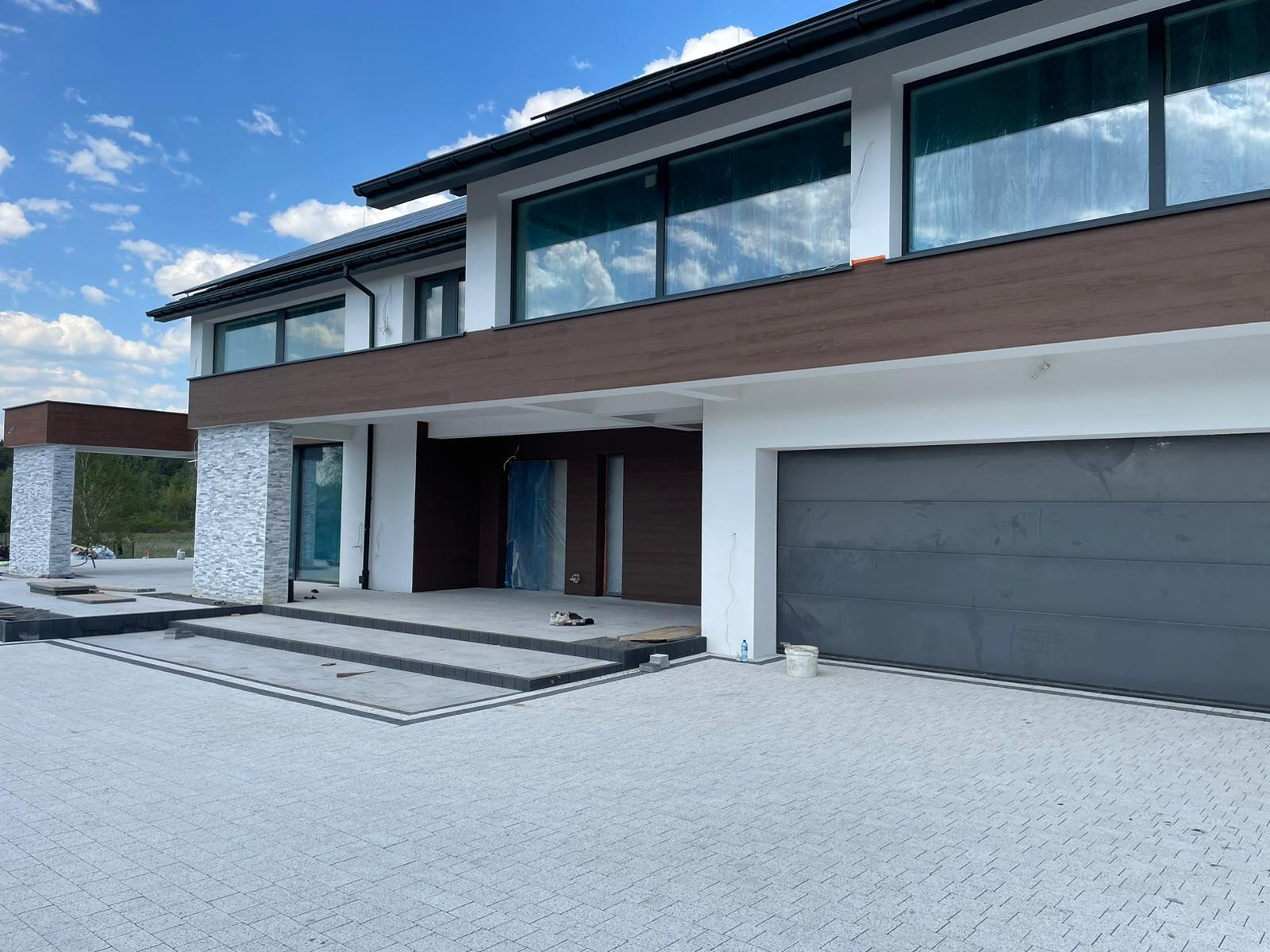 Nowoczesny dom jednorodzinny z elewacją w odcieniach bieli, brązu i szarości. Widoczne okna z odbiciem nieba i fragment nowej kostki brukowej na podjeździe.
